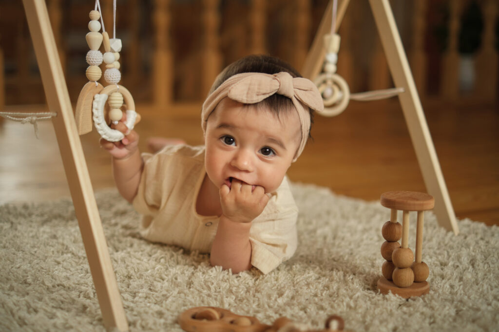 Exploring the World of Baby Teething: A one-year-old cherub lays on a beige mat, gnawing her fingers to relieve teething discomfort, surrounded by wooden toys.
