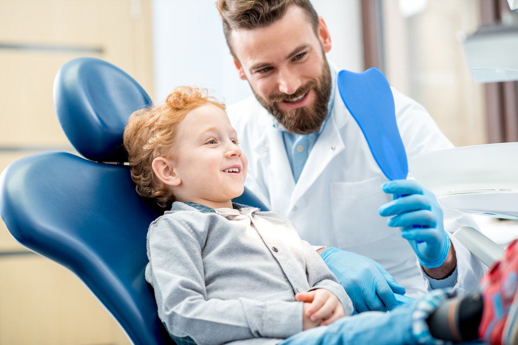Young excited boy looking at the dental mirror sitting on the chair with dentist at the dental office
