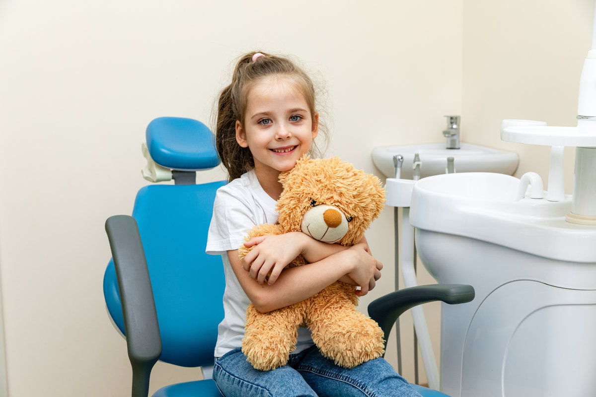 Happy little girl having dentist's appointment in modern clinic. Little patient, adorable girl sitting in dentist's chair during appointment in pediatric dentistry clinic and hugging her teddy bear.