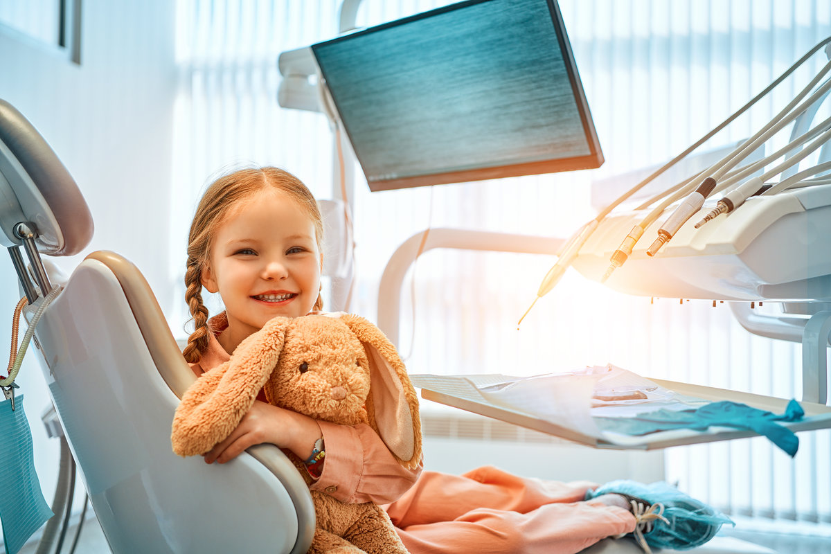 A cute little girl sitting in a dental chair, holding a toy rabbit and laughing. In the background, dental equipment and a monitor for the clinic logo. Dental care, medicine.Sunlight.