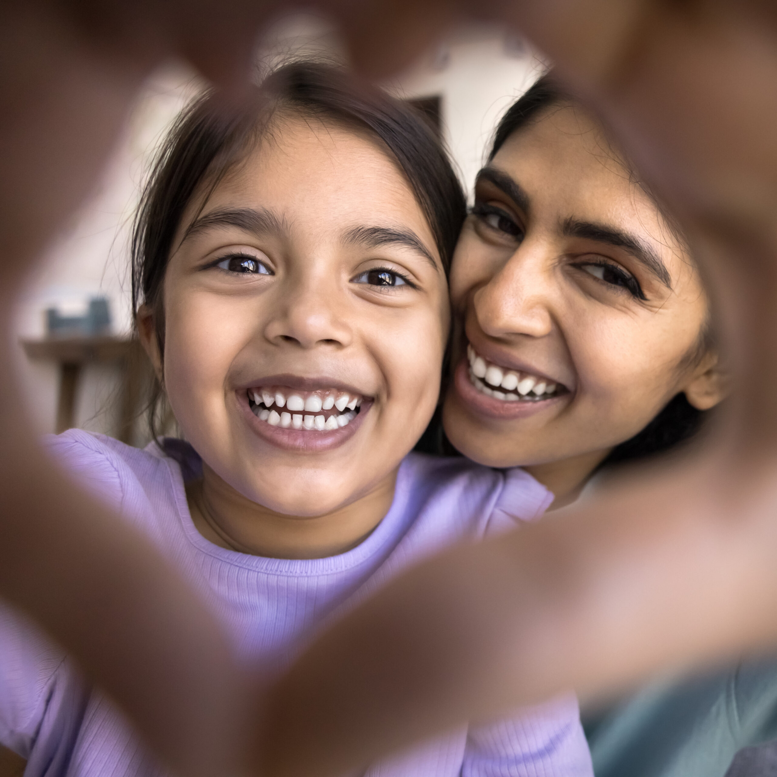 Close up faces through joined finger showing Indian woman and her adorable little 5s daughter stare at camera revealing white teeth, advertising dental clinic services. I love you, declaration of love