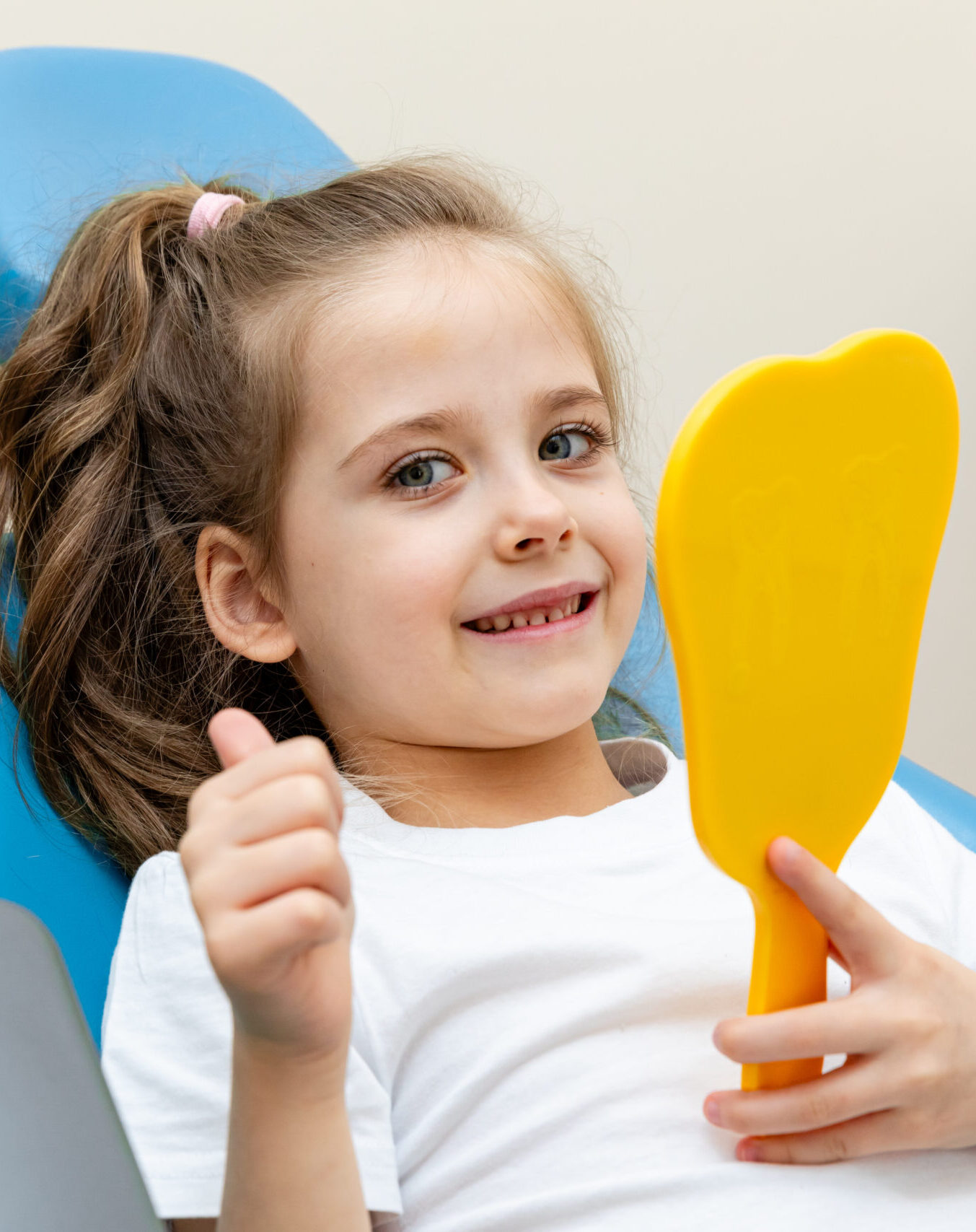 Portrait of a cute happy little girl sitting on a chair in a dental office, showing thumbs up after a dental examination while holding mirror in her hand.