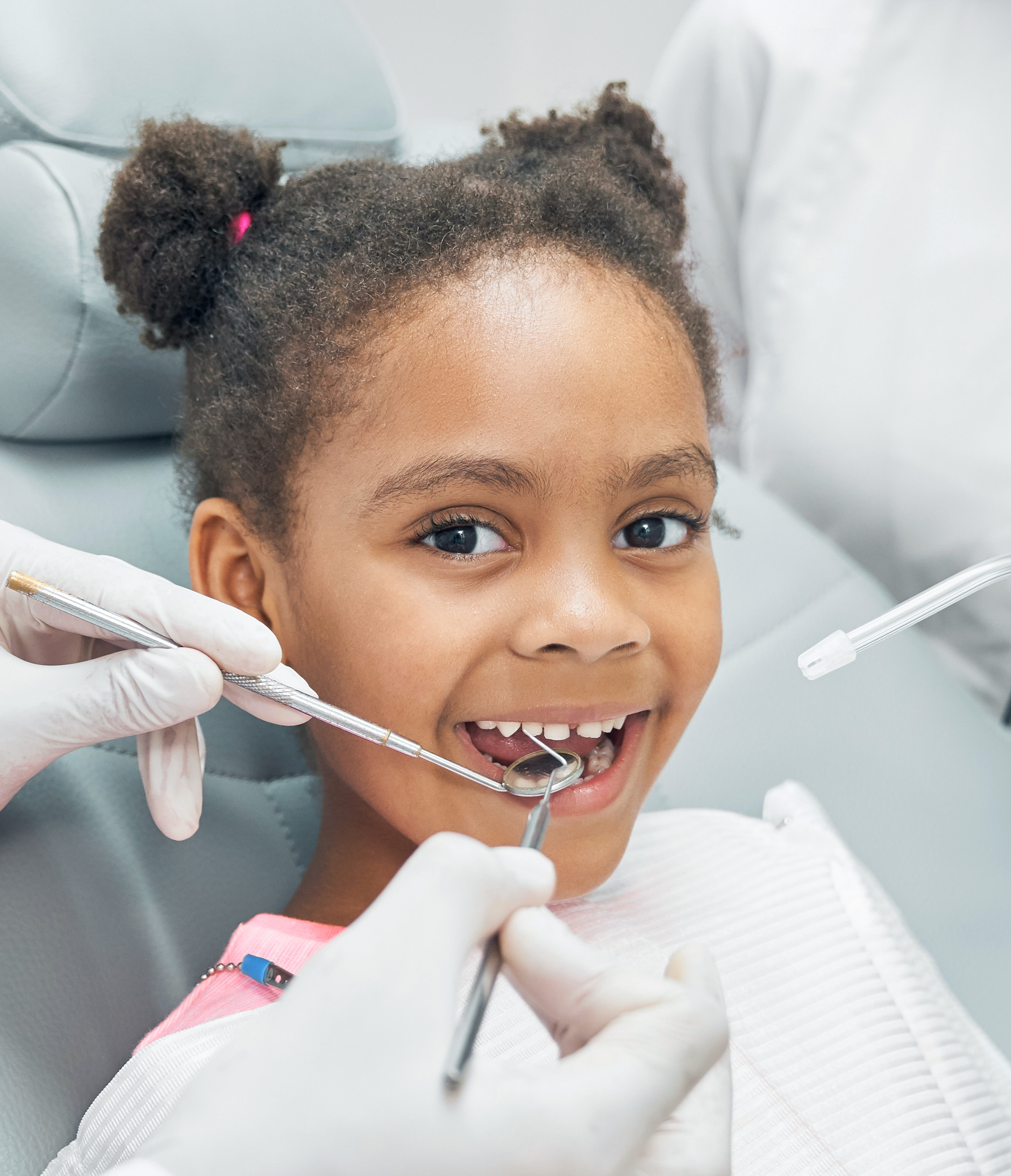Happy afro american girl sitting in stomatologist chair with open mouth while professional dentist doing regular check up of teeth using dental probe and mirror. Female nurse assisting.
