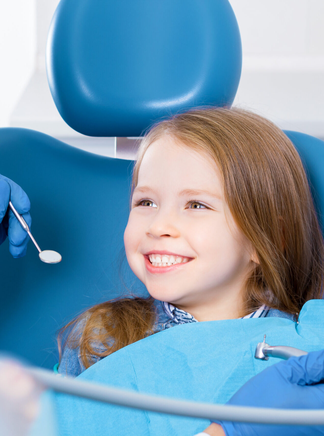 Little cute smiling girl is sitting in dental chair and opening mouth in clinic, office. Doctor is preparing for examination of child teeth with tools, instruments. Visiting dentist with children.