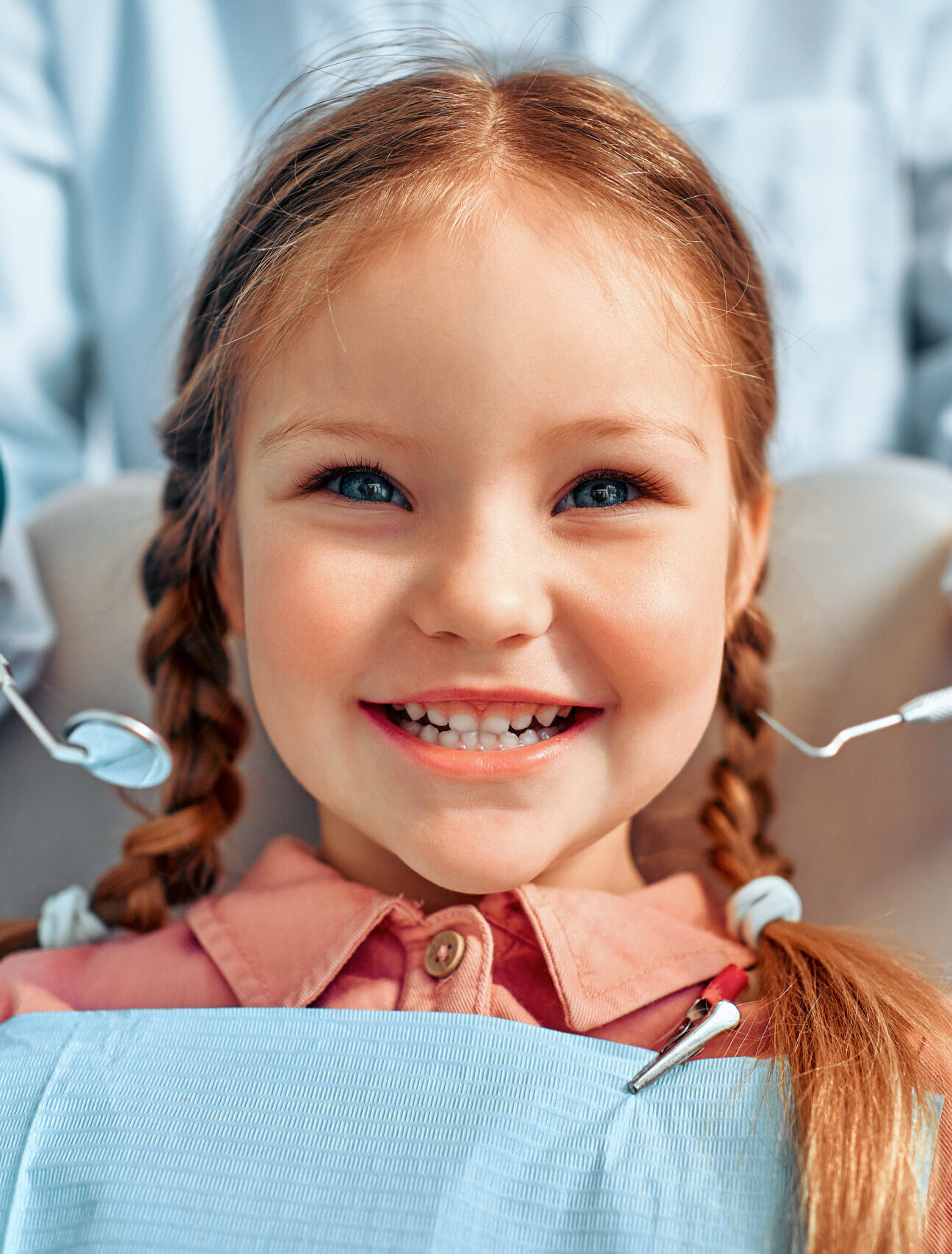 Cropped portrait of little girl with pigtails hair sitting in dental chair looking at camera and smiling. Behind, a doctor in gloves holds examination tools.Children's dentistry.