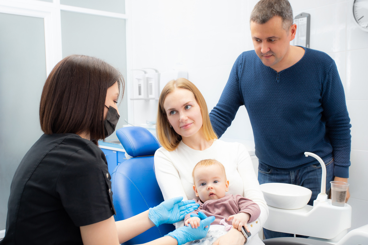 Young parents and their daughter visit a dentist to examine milk teeth. Baby at the first appointment with the dentist. Inspection of the formation of jaws, eruption of milk teeth.