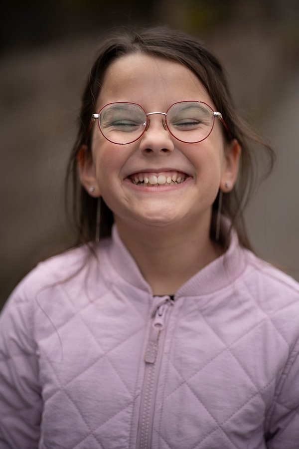 Smiling girl in glasses and a pink jacket showing her teeth outdoors, soft natural light, blurred background, copy space. Concept of dental health, confidence, and childhood joy.